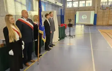 Poczet Rady Rodzic&oacute;w, przewodniczący Samorządu Uczniowskiego, wicedyrektor R. Konat podczas uroczystości. W tle na ekranie zdjęcie nowego sztandaru