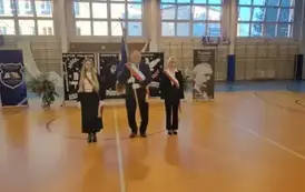 Poczet sztandarowy Rady Rodzic&oacute;w z nowym sztandarem na środku hali; w tle dekoracja z patronem szkoły i bukiety kwiat&oacute;w